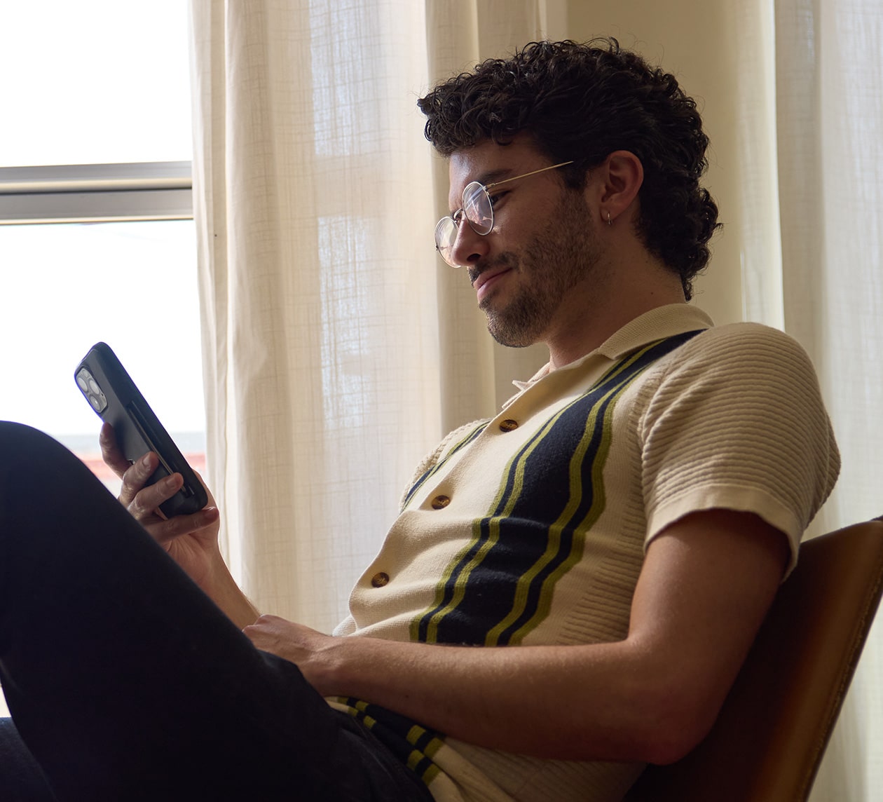 A person sits relaxed in a chair, holding a smartphone, with sunlight filtering through sheer curtains in the background.