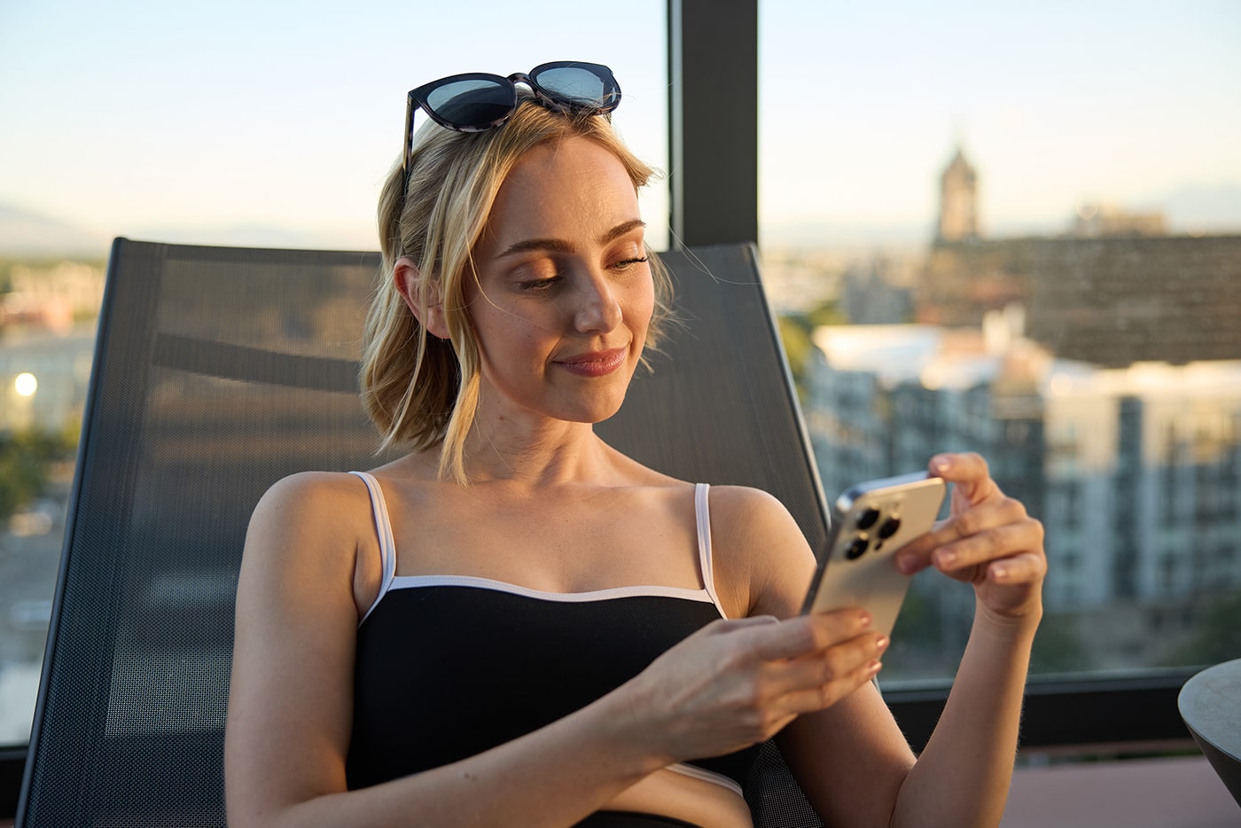 A woman in a black top relaxes on a lounge chair, using her phone against a backdrop of a cityscape at sunset.