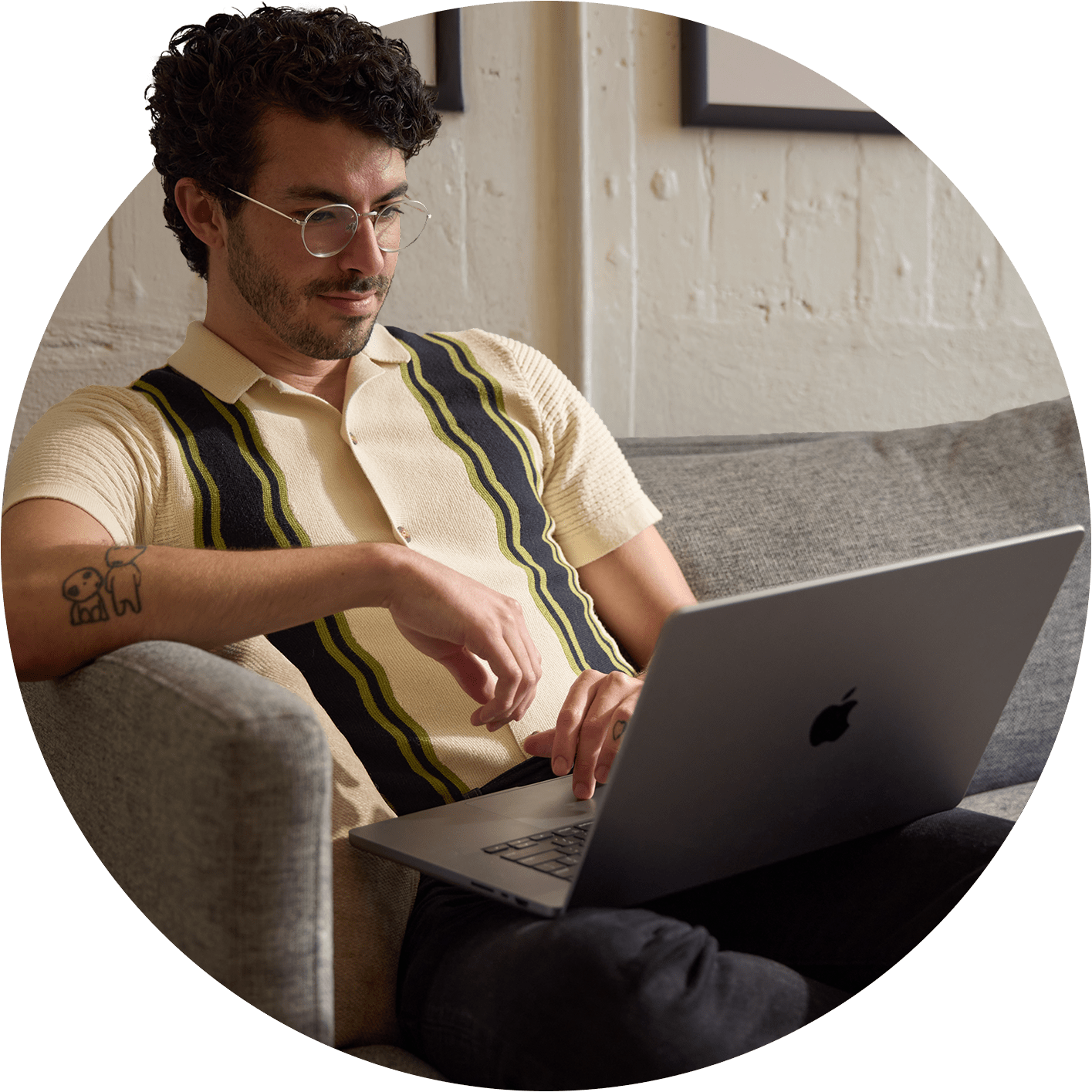 A person with curly hair sits comfortably on a gray sofa, working on a laptop while wearing a retro-style striped shirt.