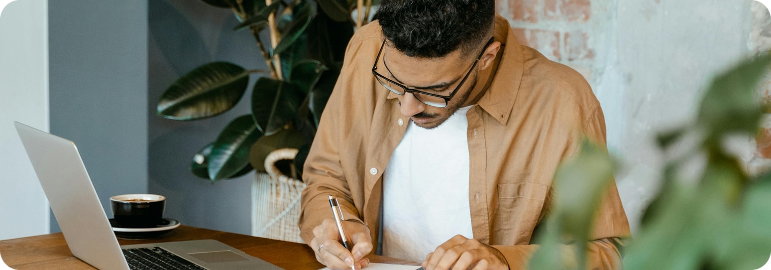 Man writing on a notepad with a laptop open beside him